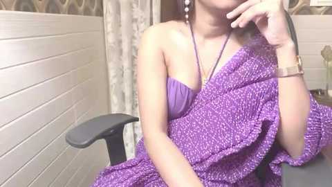 Media: Video of an Asian woman with light skin, wearing a purple, patterned saree and a gold watch, seated in an office chair against a beige wall with a grey cushion.