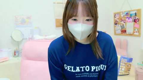 A video of a young woman with long brown hair, wearing a blue \"Gelato Pilot\" sweatshirt and a white mask, in a brightly lit, decorated room with pastel-colored walls, posters, and a pink chair.