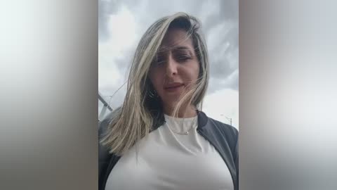 Media: A video of a woman with long, straight, blonde hair, wearing a black blazer over a white turtleneck, standing against a cloudy sky backdrop, with a soft, blurred lighting effect.