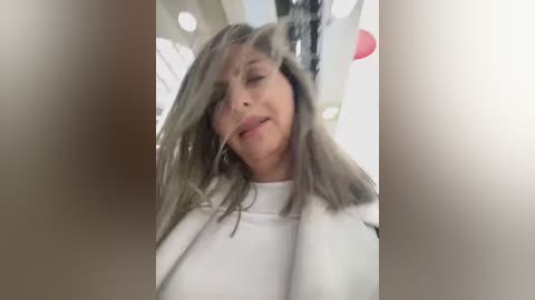 Media: A candid video of a middle-aged woman with long, gray hair, wearing a white shirt, looking upward with a neutral expression. The background is blurred, featuring an indoor setting with soft lighting.