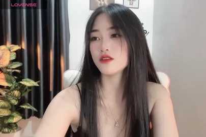 Media: Video of an East Asian woman with long black hair, light skin, and red lipstick, seated indoors. She wears a black top and necklace, with a green plant and curtains in the background.