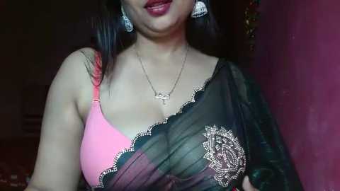 Media: Video of a South Asian woman with fair skin, wearing a pink bra, black sheer saree, and silver earrings. She has long dark hair, a gold necklace, and stands against a red wall.