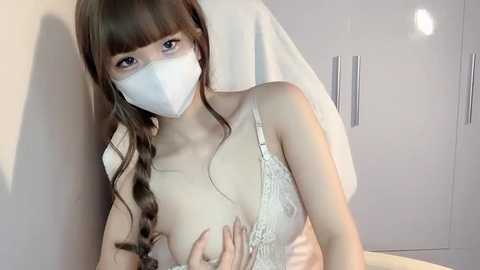 Media: A video of a slender, fair-skinned Asian woman with long brown hair, wearing a white mask and lace bra, sitting on a bed, in a minimalist room with white walls and a towel.