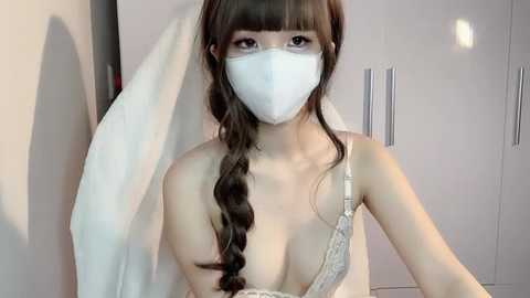 Media: Video of an Asian woman with fair skin, wearing a white surgical mask, bra, and a white towel draped over her shoulders, standing in a minimalist, light-colored room with white cabinets.