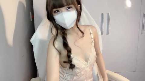 Media: Video of a young Asian woman with long brown hair, wearing a white face mask and lace lingerie, sitting on a bed in a white room with closet doors.