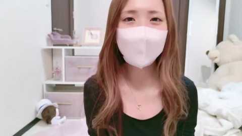 Media: A video of a young Asian woman with long, light brown hair wearing a white mask, black top, and a delicate necklace, in a softly lit bedroom with white furniture, a teddy bear, and a small white dog.