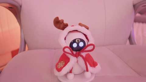 Media: Video of a plush, white, reindeer-themed doll with a black face and glowing eyes, wearing a red hooded jacket, seated on a white leather couch in a warmly lit room.