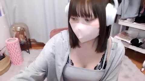 Media: Video of a young Asian woman with straight brown hair, wearing white headphones, a light blue hoodie, and a black lace bra, sitting in a cluttered room with a pink rolling pin, a helmet, and a white shelf in the background.