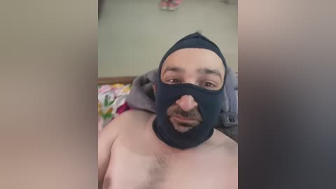 Media: A video of a fair-skinned, bearded man wearing a black mask with a prosthetic nose, standing shirtless in a bedroom with a floral-patterned bedspread.