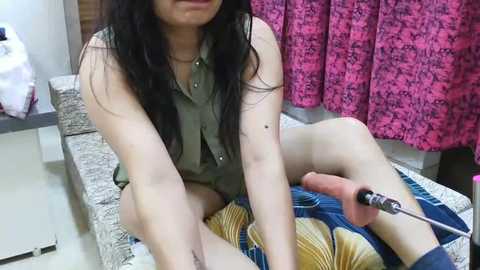 Media: Video of a woman with long black hair, wearing a sleeveless olive-green top and shorts, sitting on a patterned sofa. She is using a pliers to remove a splinter from her foot, with a pink curtain and a wooden cabinet in the background.