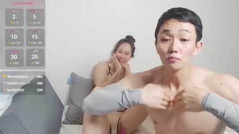 Media: A video shows an Asian woman in a gray tank top and a man with short black hair, both topless, in a bedroom. The background features a white wall and gray pillows on a bed.