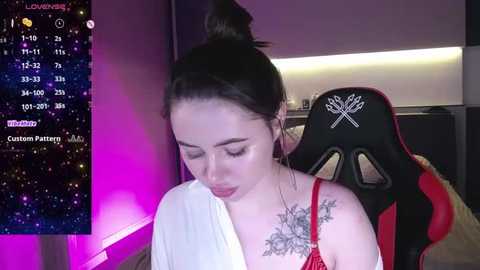 Media: Video of a fair-skinned woman with dark hair in a bun, wearing a red bra, and a white robe, sitting in a gaming chair. Background shows a dark room with a pink and purple light, a TV displaying a game, and a starry sky overlay.