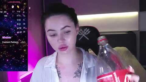 Media: Video of a young woman with fair skin and dark hair in a bun, wearing a white shirt, holding a red drink bottle. Background shows a dimly lit room with a starry screen and a black gaming chair.