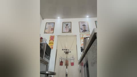 Media: Video of a narrow kitchen with white walls, featuring a beige curtain with hanging coffee mugs, framed coffee advertisements, and stainless steel appliances.