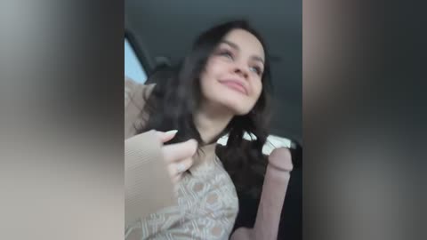 Media: A video of a young woman with fair skin and wavy dark hair, wearing a beige sweater and a brown leather seatbelt, smiling and looking up in a car.