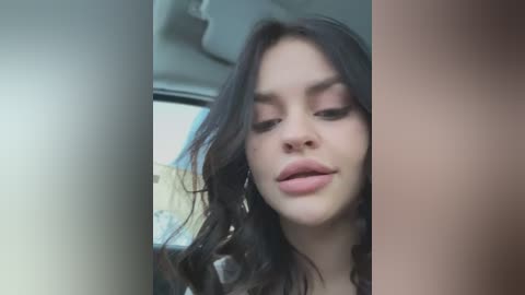 Media: A close-up video of a young woman with long, wavy dark hair, fair skin, and full lips, wearing minimal makeup. She's inside a car, with blurred background.