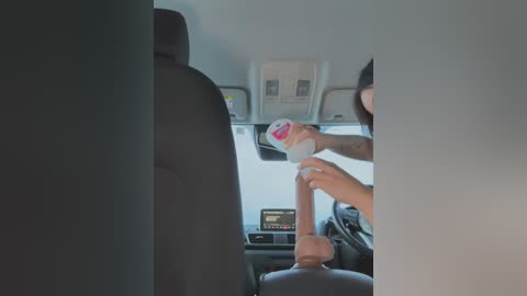 Media: Video of a car interior: a person in a black suit pours a white drink into a plastic cup held by a person in a dark jacket.