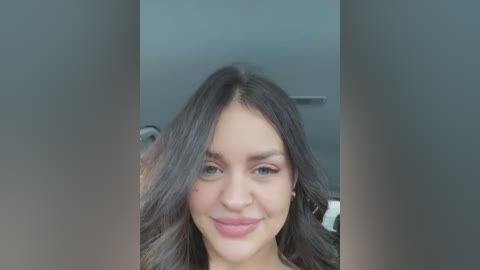 Media: A video of a young woman with wavy, dark brown hair and light skin, smiling gently. The background features a modern, gray-walled room with a partially visible mirror and a gray chair.