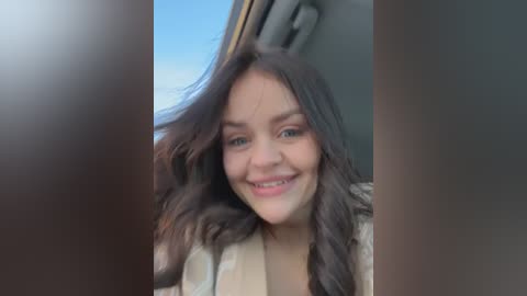 Media: Video of a young woman with long, wavy brown hair and light skin, smiling widely. She wears a beige coat and is seated in a car with a clear blue sky visible through the window.