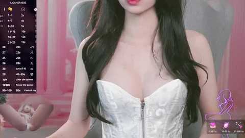 Media: Video of an Asian woman with long black hair, wearing a white corset, sitting in a modern chair. Background shows a pink wall and digital icons.