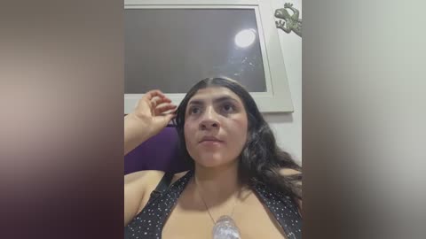 Media: Video of a woman with long dark hair, wearing a black halter top, sitting indoors. She has medium skin tone and is looking at a light source, causing a reflection in the background.