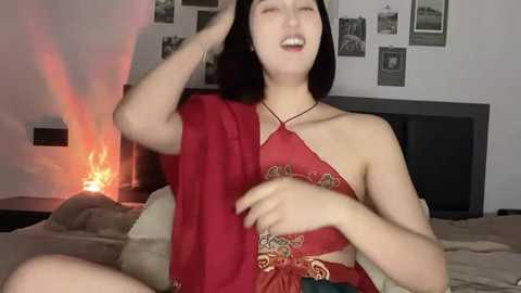 Media: Video of a smiling East Asian woman with shoulder-length black hair, wearing a red halter top, sitting on a bed with beige bedding. The background features a fireplace and framed photos on a white wall.