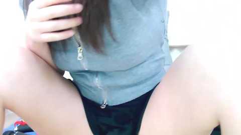 Media: A video of a woman with light skin, long brown hair, wearing a grey zip-up hoodie and black shorts, squatting with legs apart, partially covering her face with her hand.