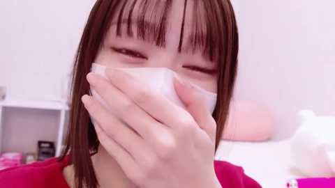 Media: Video of an East Asian woman with straight brown hair, wearing a pink shirt, holding a white tissue to her nose, indoors with a white wall and blurred background.