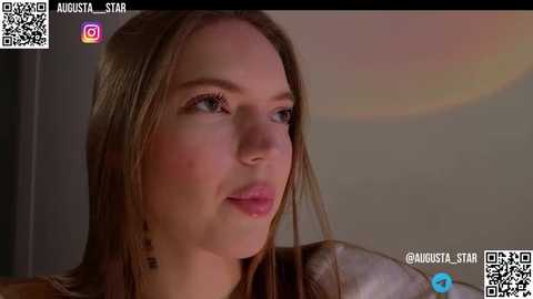 Media: Video of a fair-skinned woman with long, straight brown hair, wearing a silver necklace, looking contemplative in a soft, warm-toned room.