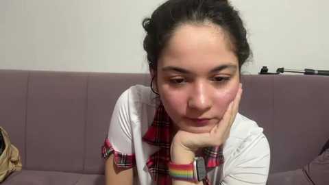 Media: Video of a young woman with light skin and dark hair, wearing a white shirt with a red plaid collar, resting her chin on her hand on a mauve couch.