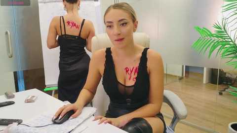 Media: Video of a blonde woman in a black lace dress, seated at a desk, with \"RYAN\" written in red on her chest. A mirror shows her back, in a modern office setting with a green plant.