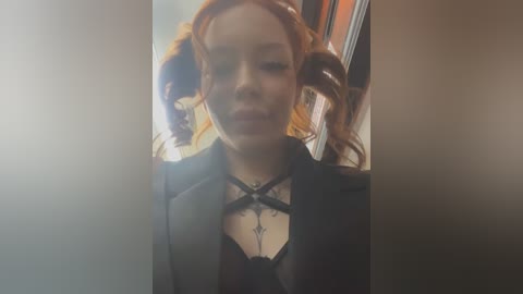 Media: A video of a young woman with pale skin, fiery red hair, and light makeup, wearing a black lace-up bodysuit and jacket, standing indoors with a blurred background.