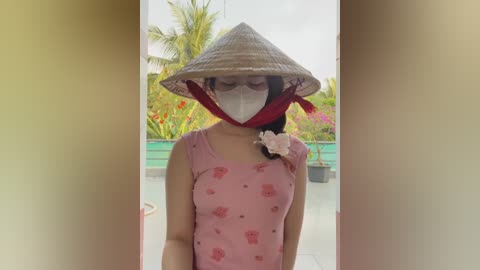 Media: Video of a woman in a pink floral dress with a conical hat, holding a flower, standing outside amidst vibrant tropical plants and a turquoise pool.