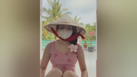 Media: Video of a smiling Asian woman with a conical hat, wearing a pink floral dress and a mask, sitting on a balcony overlooking a lush tropical garden.