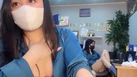 Media: Video of two women in a medical office. One woman, wearing a blue shirt, takes a selfie, showing cleavage. Another woman, masked, sits behind a desk with potted plants.