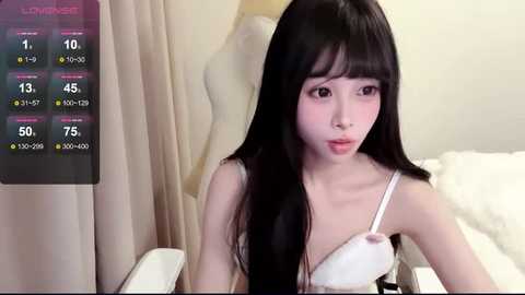 Media: Video of an Asian woman with long black hair and pale skin, wearing a white bra, sitting on a white chair in a minimalist room with beige curtains.