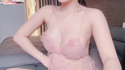 Video of an Asian woman with light skin, wearing a sheer, pink tulle dress with ruffled edges, revealing her medium-sized breasts. She has a delicate necklace and sits on a gray couch in a modern living room.