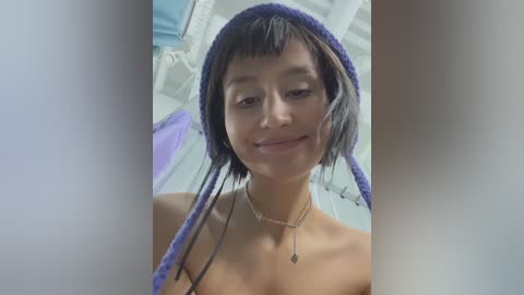 Media: Video of a young, slender, light-skinned Asian woman with straight black hair, wearing a purple crochet hat and a silver necklace, smiling slightly, standing indoors.