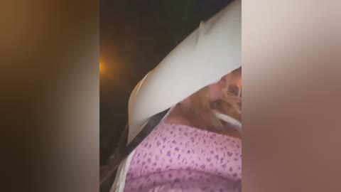 Media: A blurred video of a man with a mustache, wearing a pink hospital gown with a pattern, inside a car, with the window down, at night.