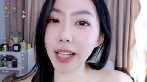 Media: A close-up video of a smiling East Asian woman with long black hair, fair skin, and subtle makeup. She wears a black top. Background includes a beige curtain and a cluttered shelf.