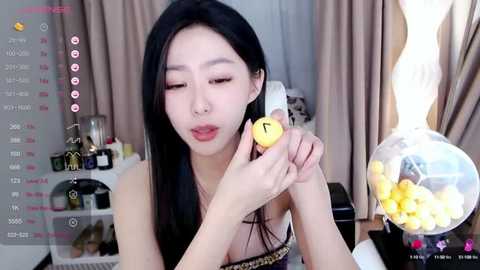 Media: Video of an East Asian woman with long black hair, light skin, and a slim physique, holding a yellow banana with a number 7 sticker, in a cozy room with beige curtains and yellow candy.