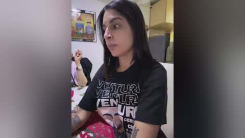 Media: Video of a woman with medium skin tone, long dark hair, wearing a black \"Venom\" T-shirt, sitting on a bed in a small, cluttered room with a TV showing a cartoon.