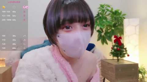 Media: Video of an East Asian woman with short, dark hair and a white mask, wearing a pink robe, in a cozy indoor setting with a Christmas tree and potted plant.