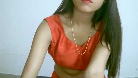 Media: Video of a young Asian woman with long, straight black hair, wearing a sleeveless, orange satin crop top with a gold necklace. She has a slender build and fair skin. The background is plain white.