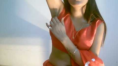 Media: Video of a light-skinned woman with long brown hair, wearing a shiny red satin top, with a tattoo on her left wrist, sitting against a plain white wall, arm raised.