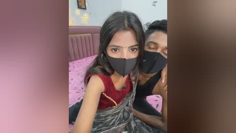 Media: Video of two women wearing black masks, sitting on a pink bed with floral patterns, one in a red blouse, the other in a black top, in a dimly lit room with yellow wall art.