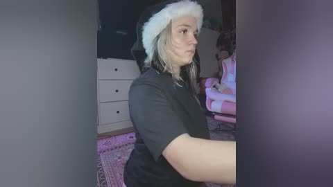 Media: Video of a young woman with long, wavy, blonde hair, wearing a black t-shirt and a festive Santa hat, standing indoors in a dimly lit room with a pink rug and white furniture.