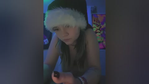 Media: Video of a young woman with fair skin and long dark hair, wearing a Santa hat, leaning forward, topless, in a dimly lit room with psychedelic posters.