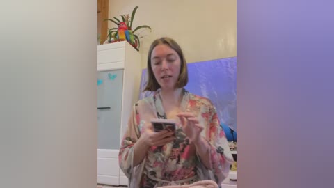 Media: Video of a woman in a floral robe, holding a smartphone, in a brightly lit room with a colorful toucan toy, white cabinets, and a blue TV screen.