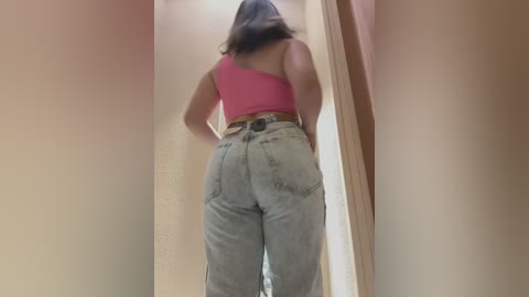 Media: Video of a woman viewed from behind, showcasing her curvy physique and large buttocks in high-waisted light-wash jeans. She wears a pink tank top and has long, dark hair.